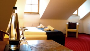 Habitación de hotel con cama, una silla, una mesa con una botella y un vaso, y una ventana con luz natural.