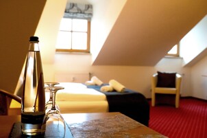 Habitación de hotel con cama, una silla, una mesa con una botella y un vaso, y una ventana con luz natural.