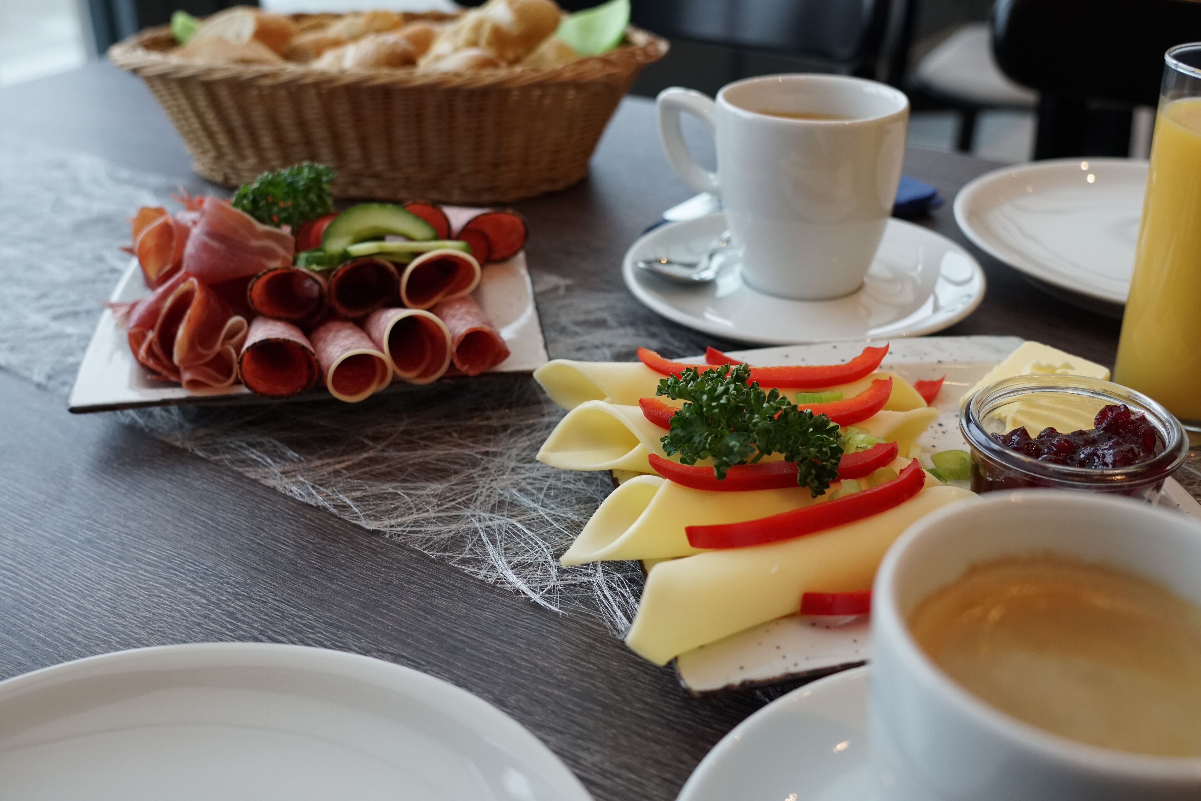 Uma mesa de café da manhã com diversos frios, queijos e legumes em um prato, uma xícara de café e uma cesta de pão.