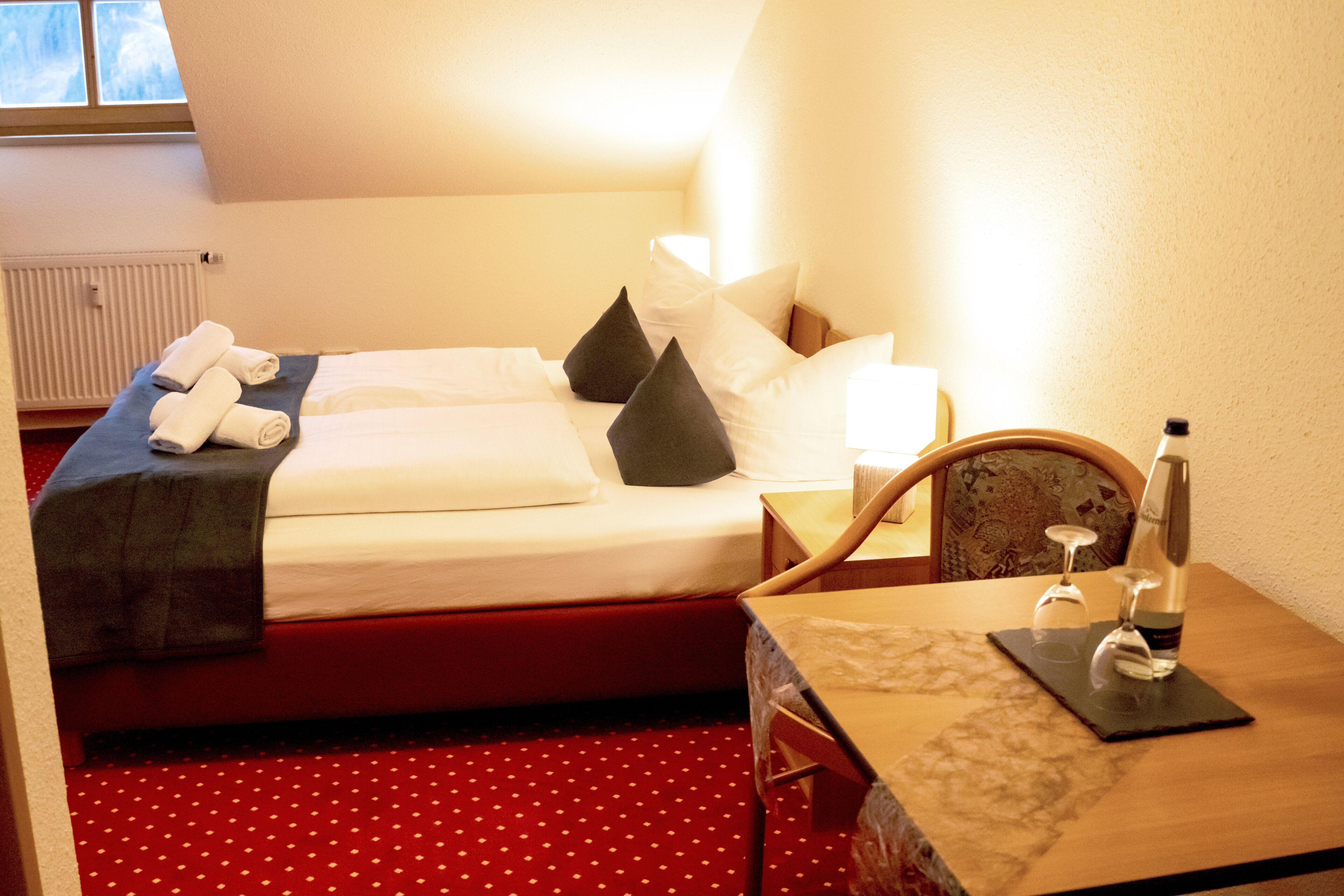 A hotel room with a bed, a table with a bottle and glasses, and a chair.