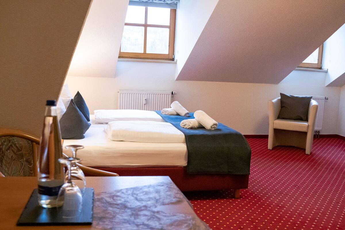 A hotel room with a bed, a chair, a table with a bottle and glasses, and a window.