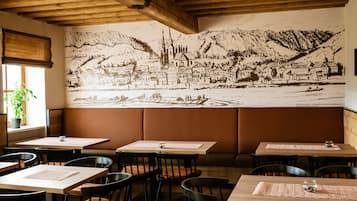 A restaurant with wooden tables and chairs, a mural of a town on the wall, and a window with a plant.