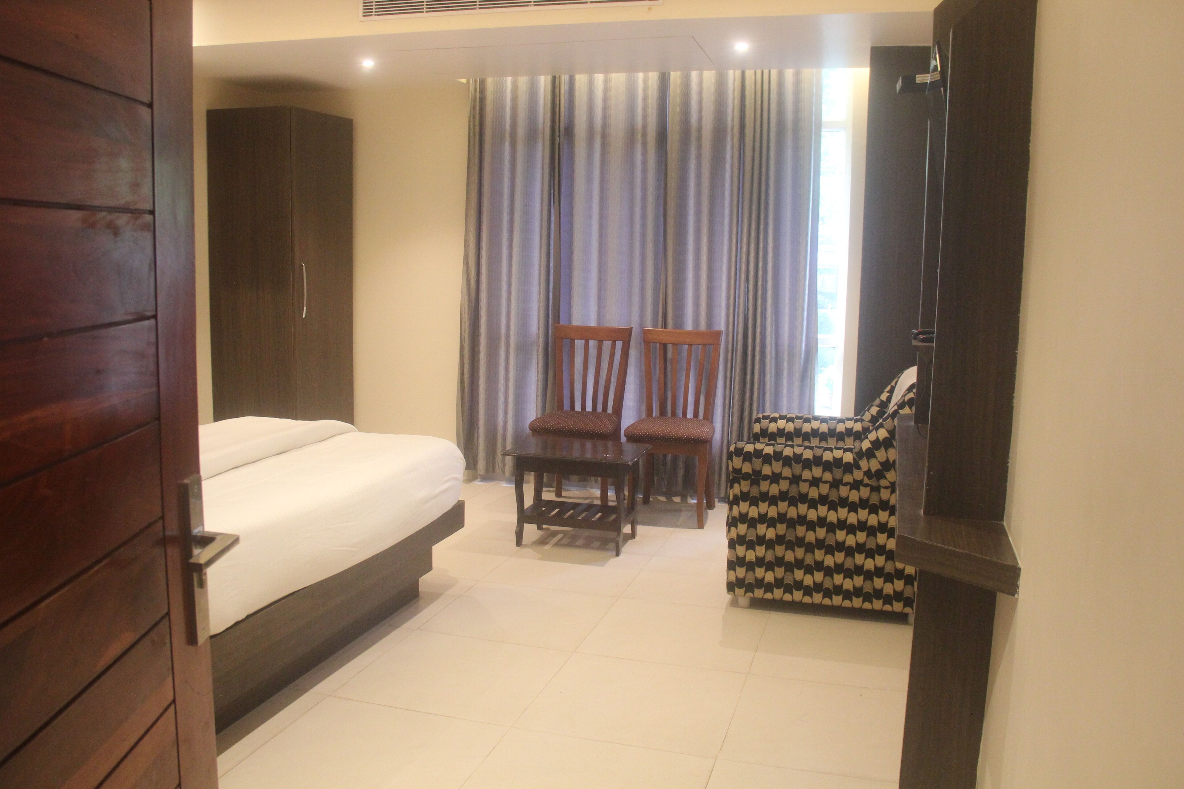 A hotel room with a bed, two wooden chairs, a small table, and a patterned armchair.