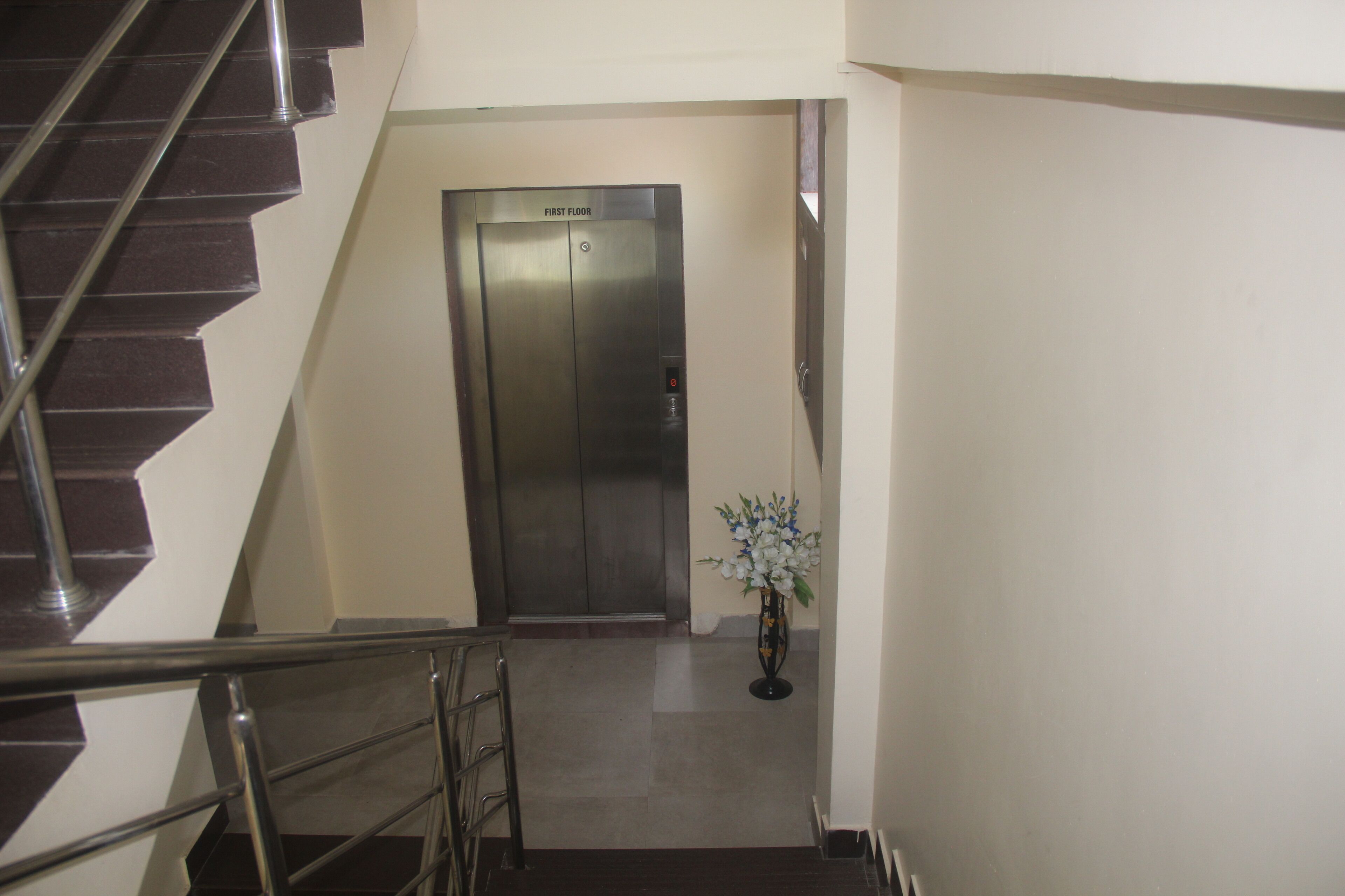 Um elevador no primeiro andar com acabamento metálico, uma escada com corrimão de metal e um vaso de plantas com flores brancas.
