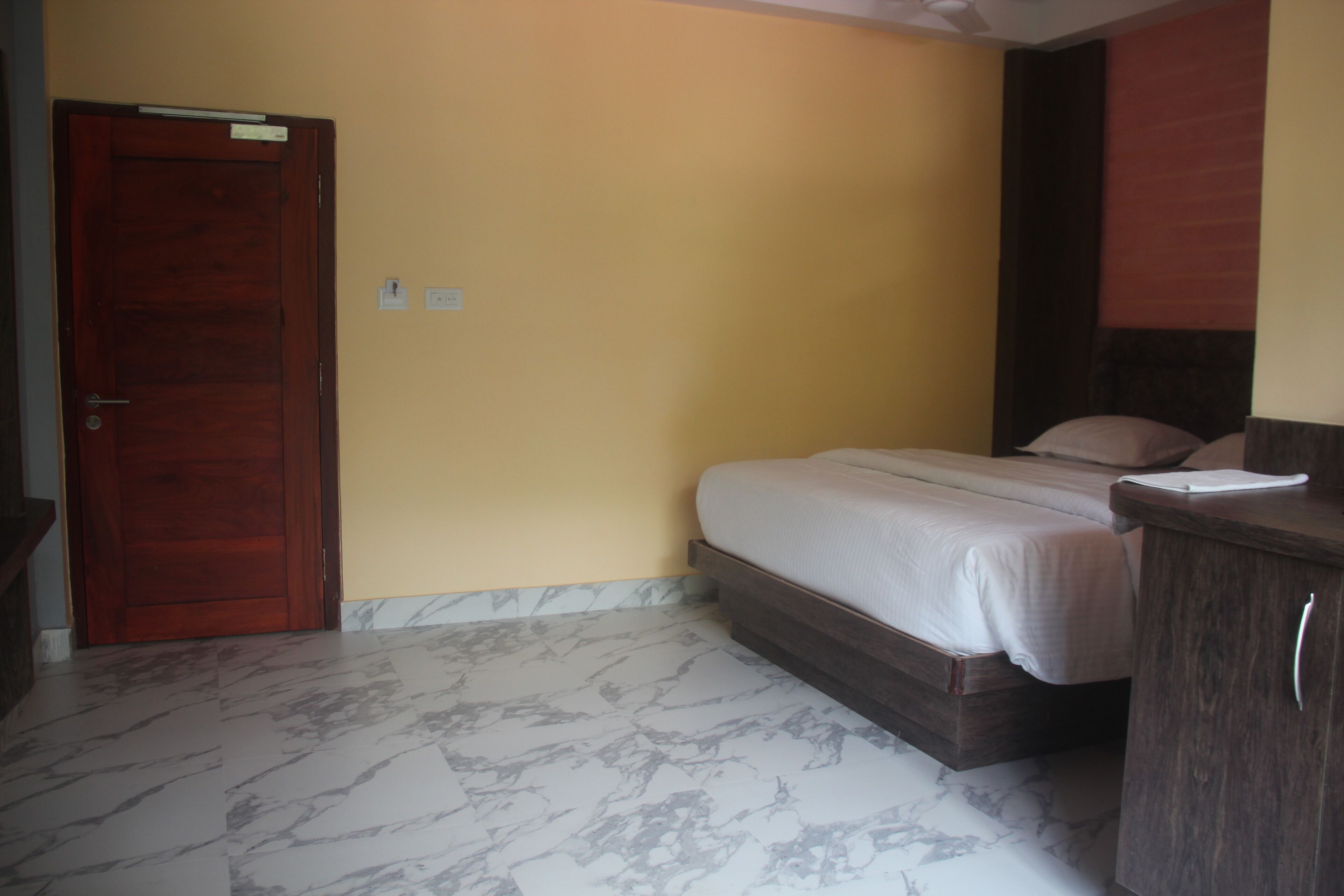 A bedroom with a wooden door, a bed with a mattress and pillows, a wooden dresser, and a marble-patterned floor.