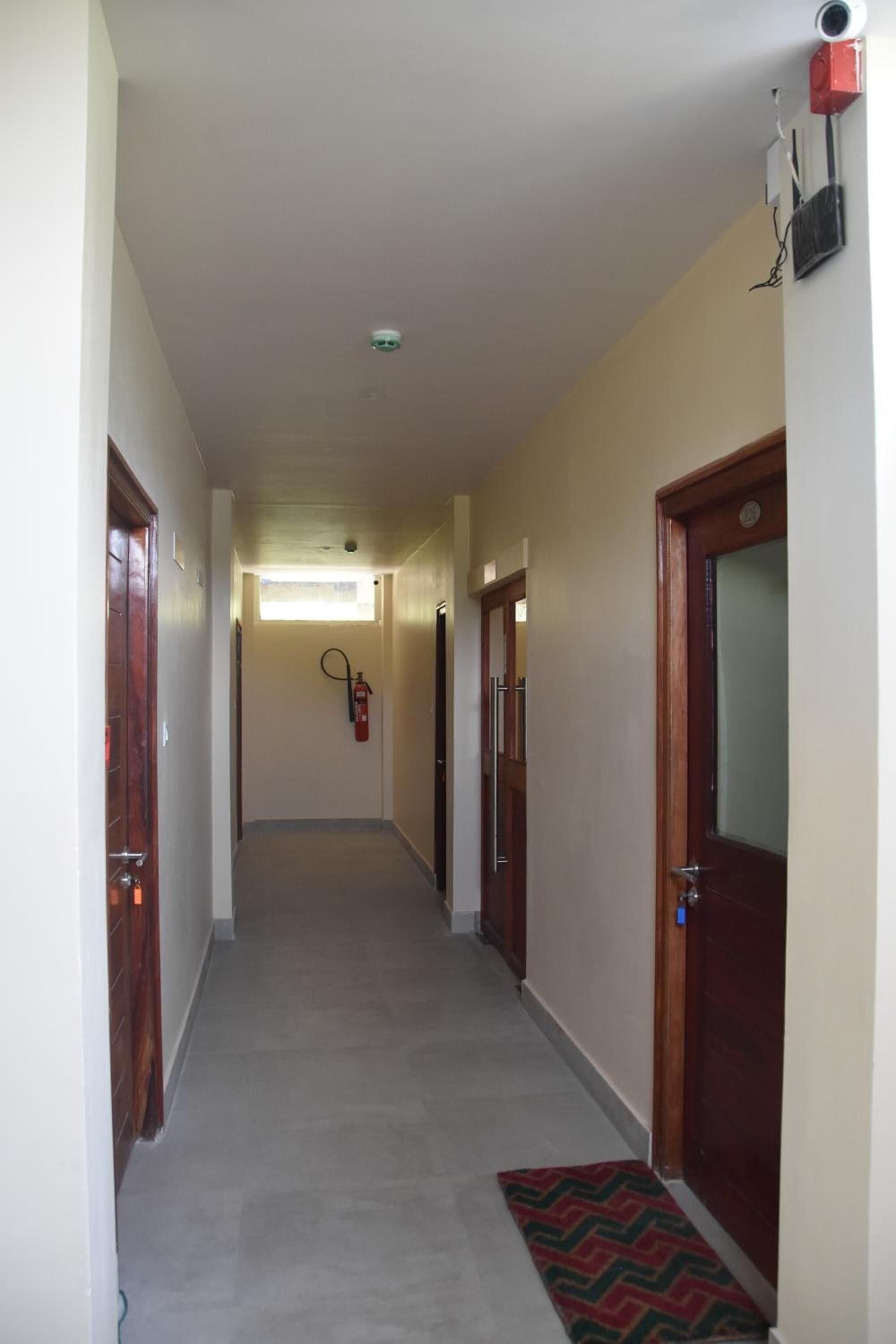 Un pasillo estrecho con puertas de madera, un extintor y una cámara de seguridad.