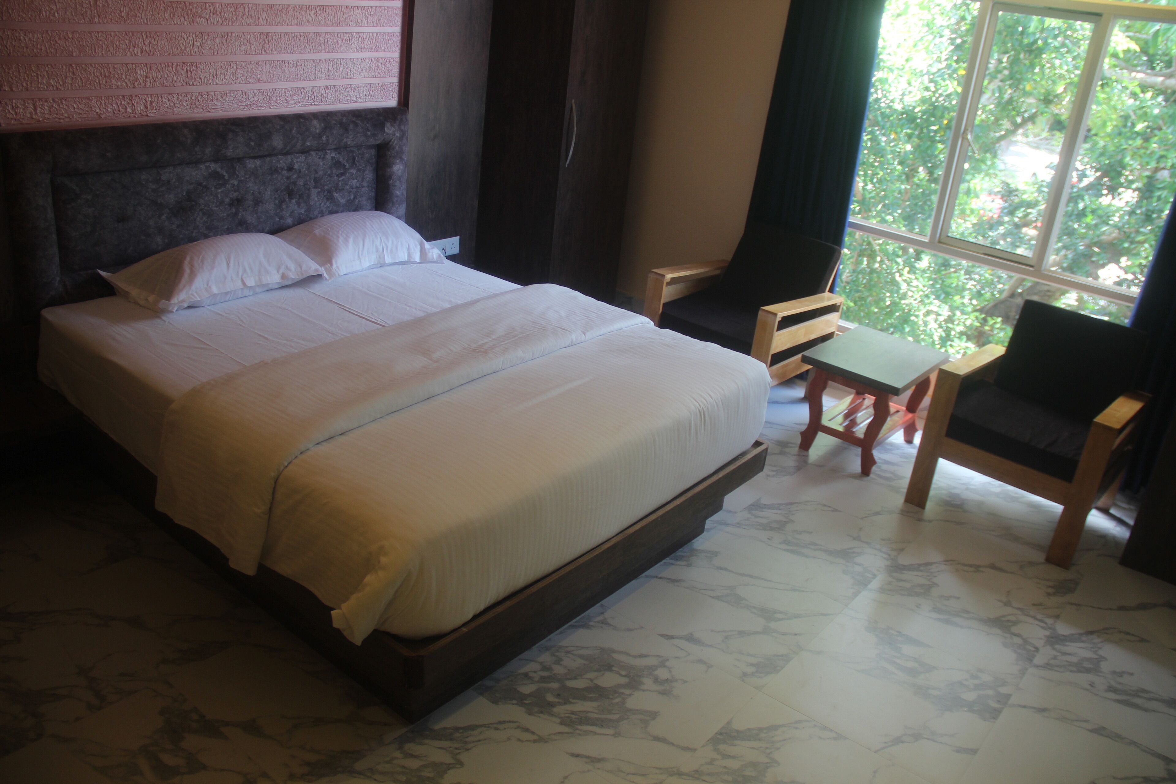 A bed with a headboard, a small table, a chair, and a window with a view of trees.