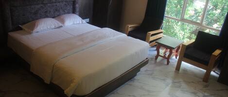 A bed with a headboard, a small table, a chair, and a window with a view of trees.