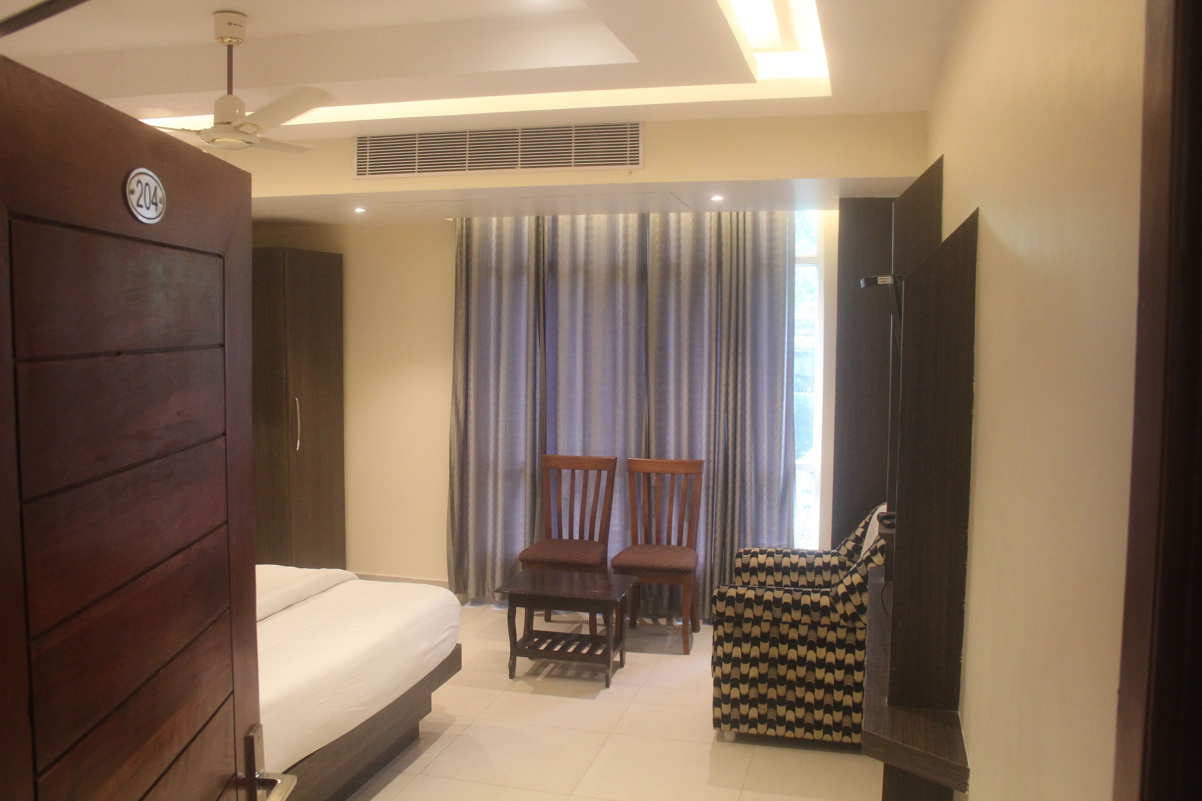 A hotel room with a bed, two chairs, a small table, a TV, and a large window with curtains.