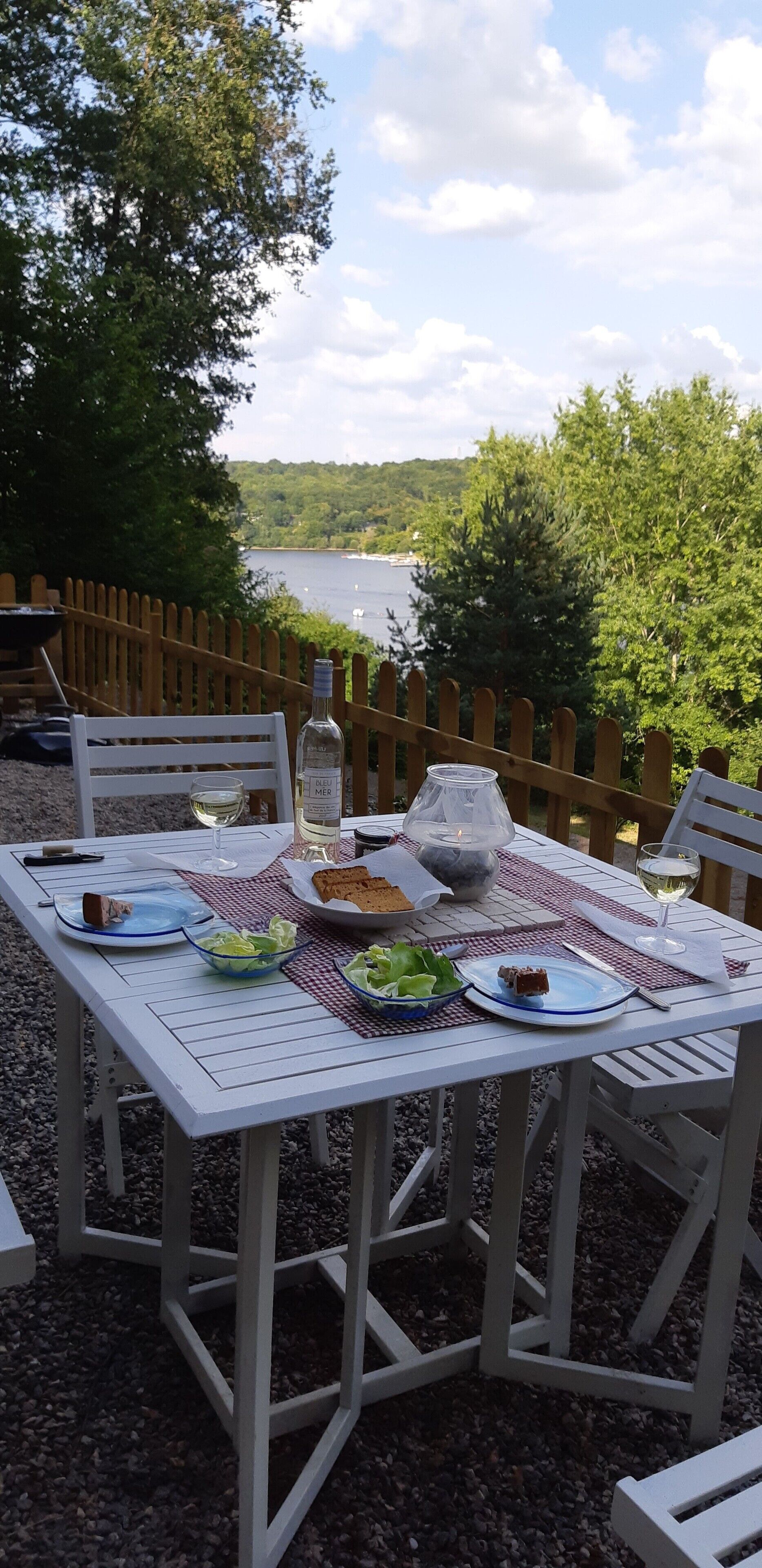 A table set outdoors with plates, glasses, and a bottle, overlooking a lake.