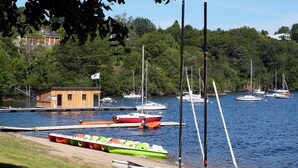 A marina with boats docked, a small wooden building, and a forested hillside in the background. - Lake view Lodge chambon Eguzon  <br>Beautiful  Lake Views  (Éguzon-Chantôme)