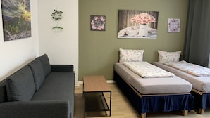 A modern, well-lit room with a grey sofa, two single beds with floral bedding, a small wooden table, and several wall decorations including a sunset painting, a floral wreath, and a couple of framed pictures.