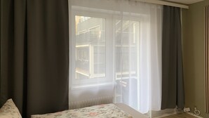 A bedroom with a bed, a window with curtains, a radiator, and a wall socket.