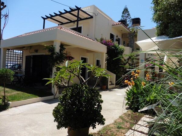 A two-story house with a covered patio, potted plants, and a small garden area. - Julie's studio,2 minutes walk to beach, near Heraklion,Knossos and famous sites! (Kokkini Hani)