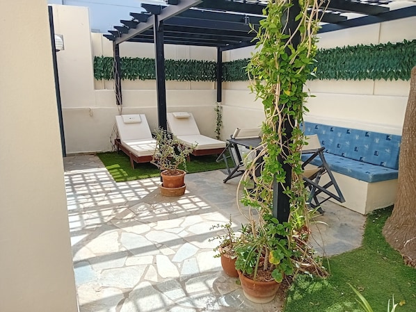 Eine Terrassenfläche mit einer Pergola, zwei Liegestühlen, einer blauen gepolsterten Bank und Kübelpflanzen.