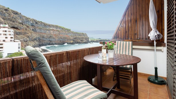 Un balcon avec une table et des chaises en bois, une plante en pot et une bouteille de vin. Le balcon donne sur une côte rocheuse, avec un bâtiment visible au loin.