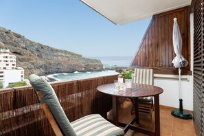Un balcon avec une table et des chaises en bois, une plante en pot et une bouteille de vin. Le balcon donne sur une côte rocheuse, avec un bâtiment visible au loin.