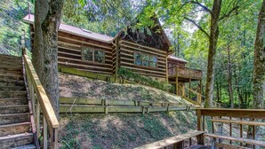 Un chalet rustique avec un escalier en bois menant à une terrasse, le tout entouré d’une forêt dense.