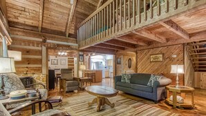 L’intérieur de cette vaste cabane en rondins est doté d’un plafond haut, d’un coin repas, d’un salon avec un canapé et des fauteuils, et d’une cuisine visible à travers une porte ouverte.