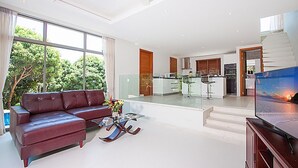 Ett rymligt vardagsrum med en modern soffa, ett soffbord i glas och en platt-TV. I bakgrunden syns ett kök med en bardisk och en trappa som leder upp till en övre våning.