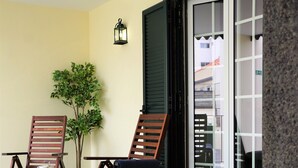 Un balcon avec deux chaises en bois, une petite table avec une boisson et des croissants, une plante en pot et un luminaire fixé au mur.