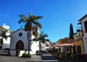 A street with a white church, palm trees, and outdoor seating areas.