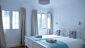 A bedroom with two beds, a window with curtains, a bedside table with a lamp, and a headboard with decorative pillows. - Apartment B - Heritage Apartments (Funchal)