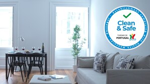 A well-lit living room with a sofa, a coffee table, and a dining area with chairs and a table. There is a window with a view of buildings outside. - Apartment B - Heritage Apartments (Funchal)