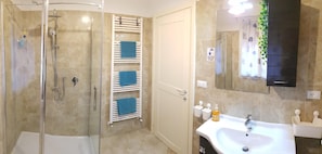A bathroom with a glass-enclosed shower, a towel rack with blue towels, a sink with toiletries, and a mirror with a plant. - B&B Corticella 24 (Bologna)