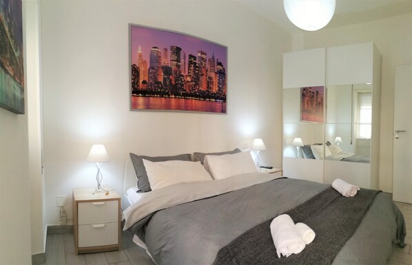 A neatly made bed with a gray blanket and two pillows, a bedside table with a lamp, a large mirror, and a cityscape painting on the wall.