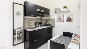 A compact kitchen with dark cabinets, a microwave, and a gas stove. There is a dining area with a table and chairs, and a small shelf with decorative items. - Nicla's Home - Near Trastevere Station (Roma)