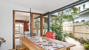 A wooden table with a floral tablecloth, a red chair, and a glass door leading to a patio with plants and a wooden fence. - Beach Cottage Lennox Head (Lennox Head)