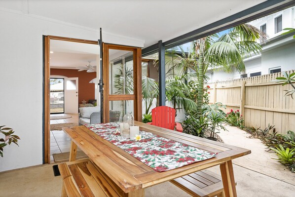 Una mesa de madera con mantel floral, una silla roja y un vidrio que da a un patio con plantas y una valla de madera.