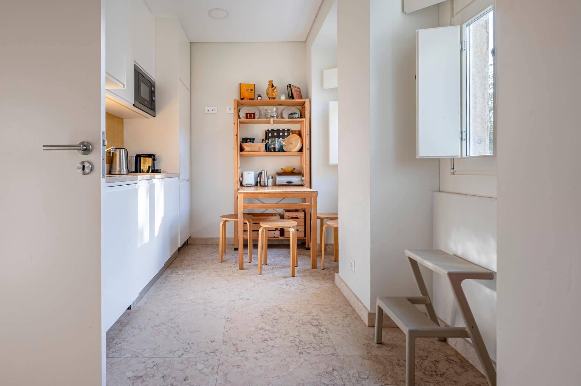 A narrow kitchen with a wooden table and chairs, a built-in shelf with kitchenware, and a window with a view.