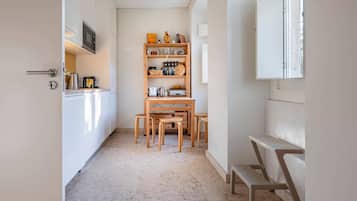 A narrow kitchen with a wooden table and chairs, a built-in shelf with kitchenware, and a window with a view.