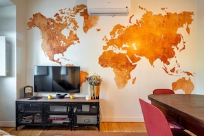 A living room with a world map mural, a flat-screen TV, a clock, a small plant, and a dining table with chairs.