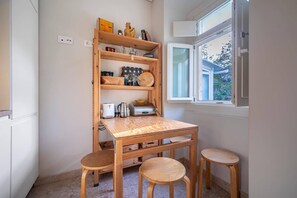A compact kitchen with a wooden table and stools, a built-in shelf with kitchenware, and a window with a view of the outside.