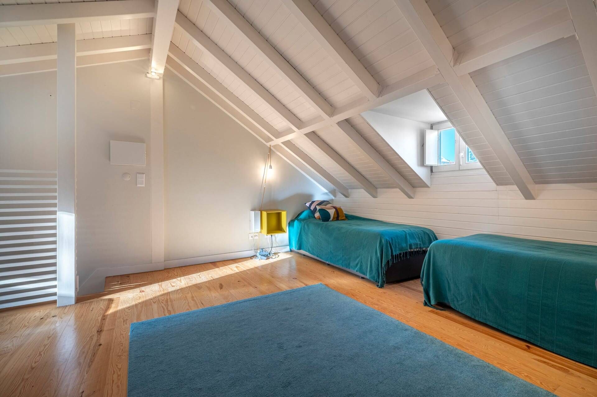 Une pièce avec un plafond en pente, un plancher en bois, deux lits avec une literie de couleur turquoise, une table de chevet jaune et une fenêtre laissant entrer la lumière du jour.