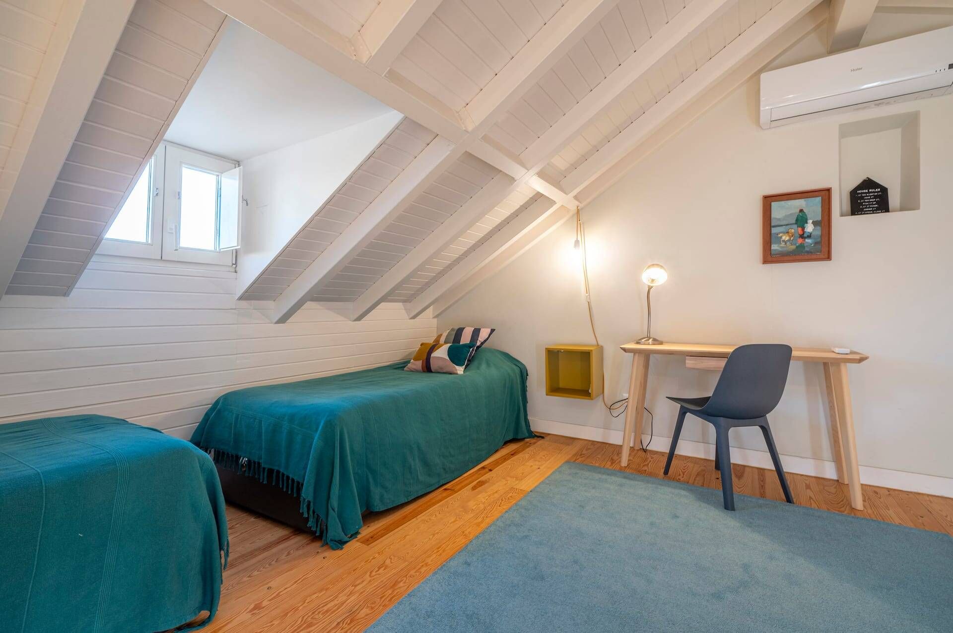 A room with a sloped ceiling, a bed with a teal bedspread, a desk with a chair, and a small window.
