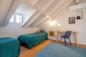 Una habitación con techo inclinado, una cama con colcha verde azulada, un escritorio con silla y una ventana pequeña.