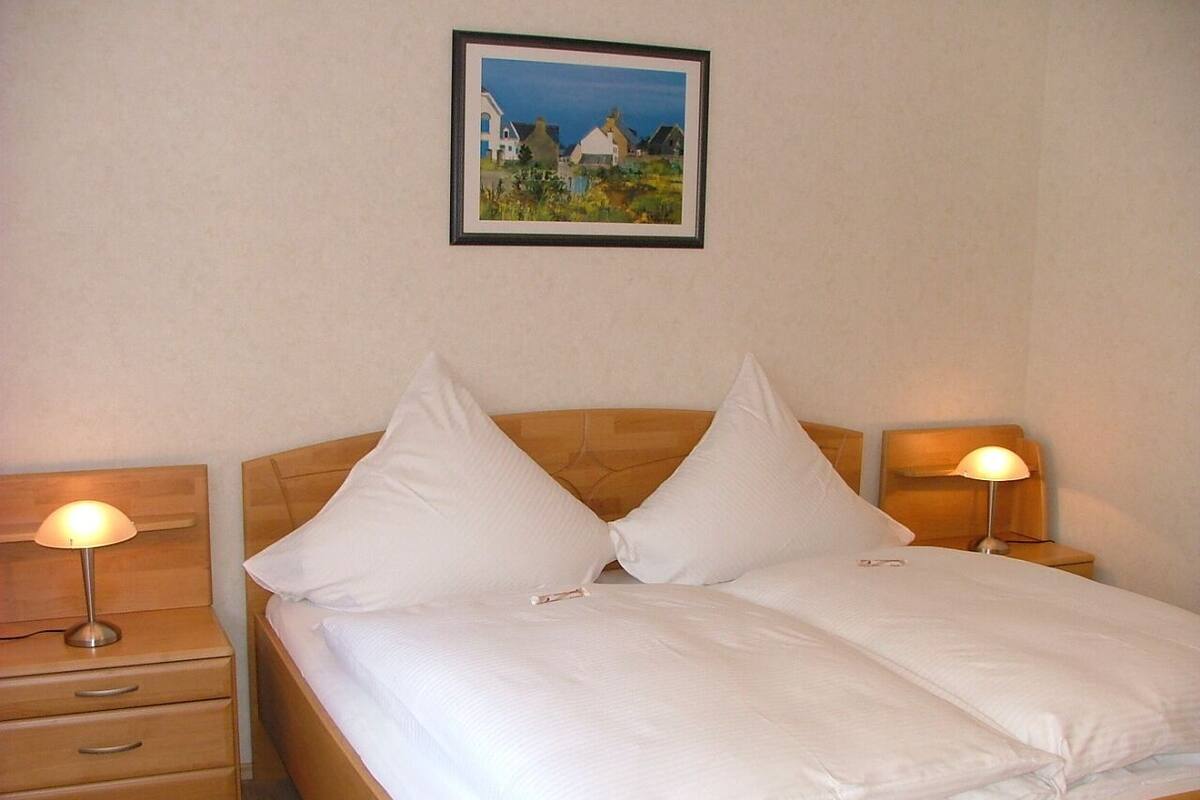 A neatly made double bed with white linens, two bedside lamps, and a framed picture on the wall.