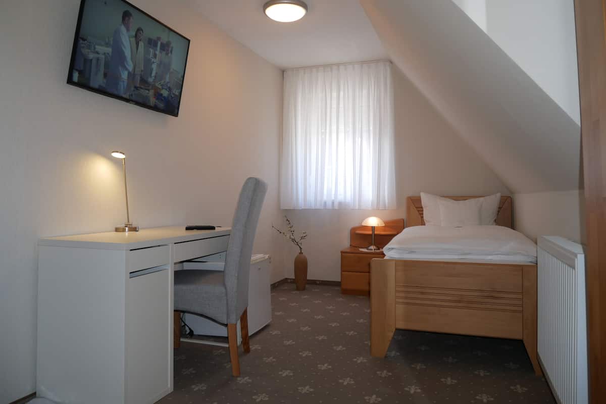 A small, well-lit room with a desk, chair, and bed. There is a painting on the wall, a window with curtains, and a nightstand with a lamp.
