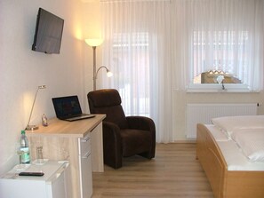 Ein Hotelzimmer mit Bett, Schreibtisch mit Laptop, Stuhl, an der Wand montiertem Fernseher und einem Fenster mit Vorhängen.