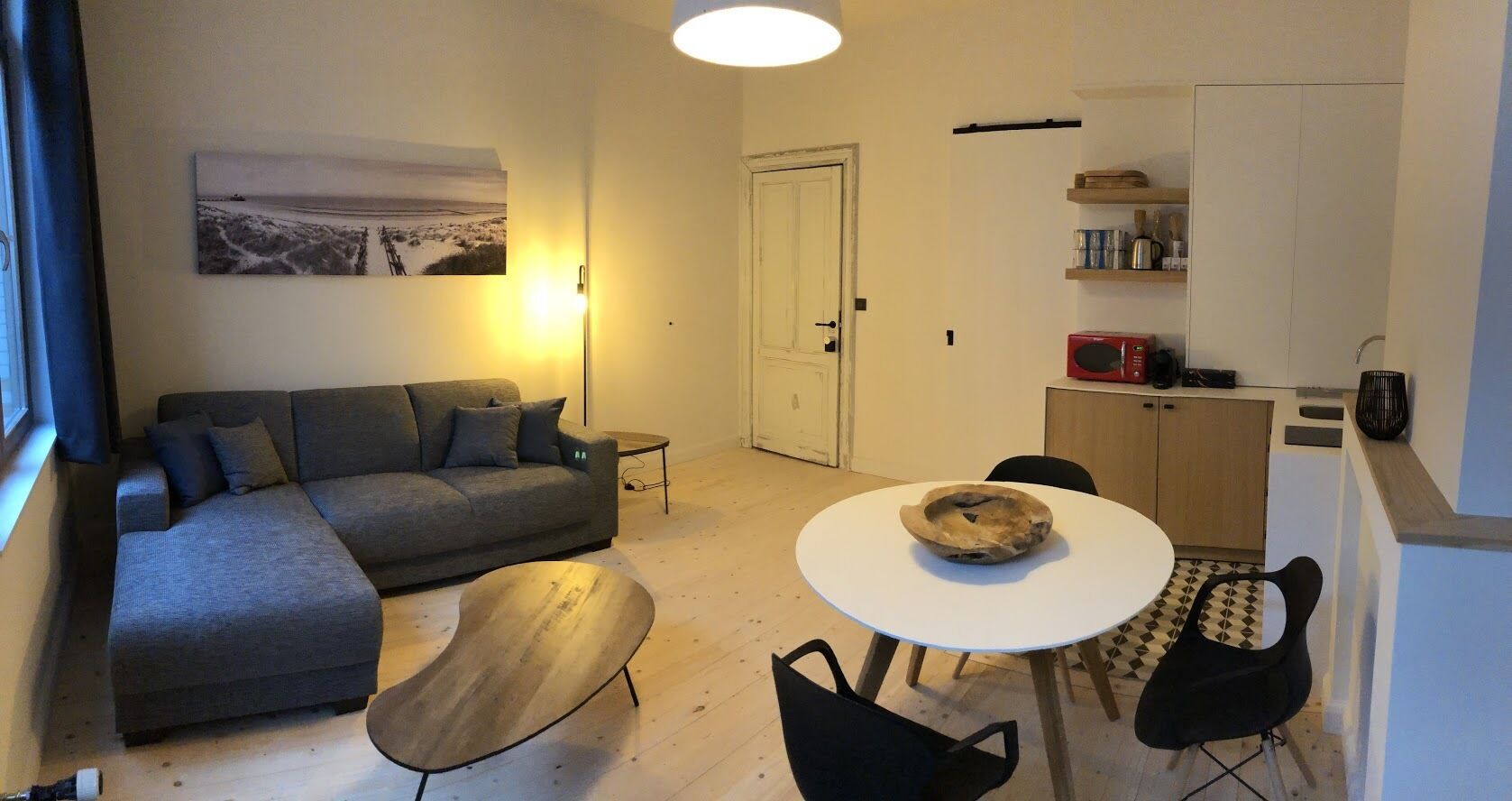 A compact living space with a grey sofa, a round wooden coffee table, a white dining table with a bowl, a black chair, and a kitchen area with a microwave and shelves.