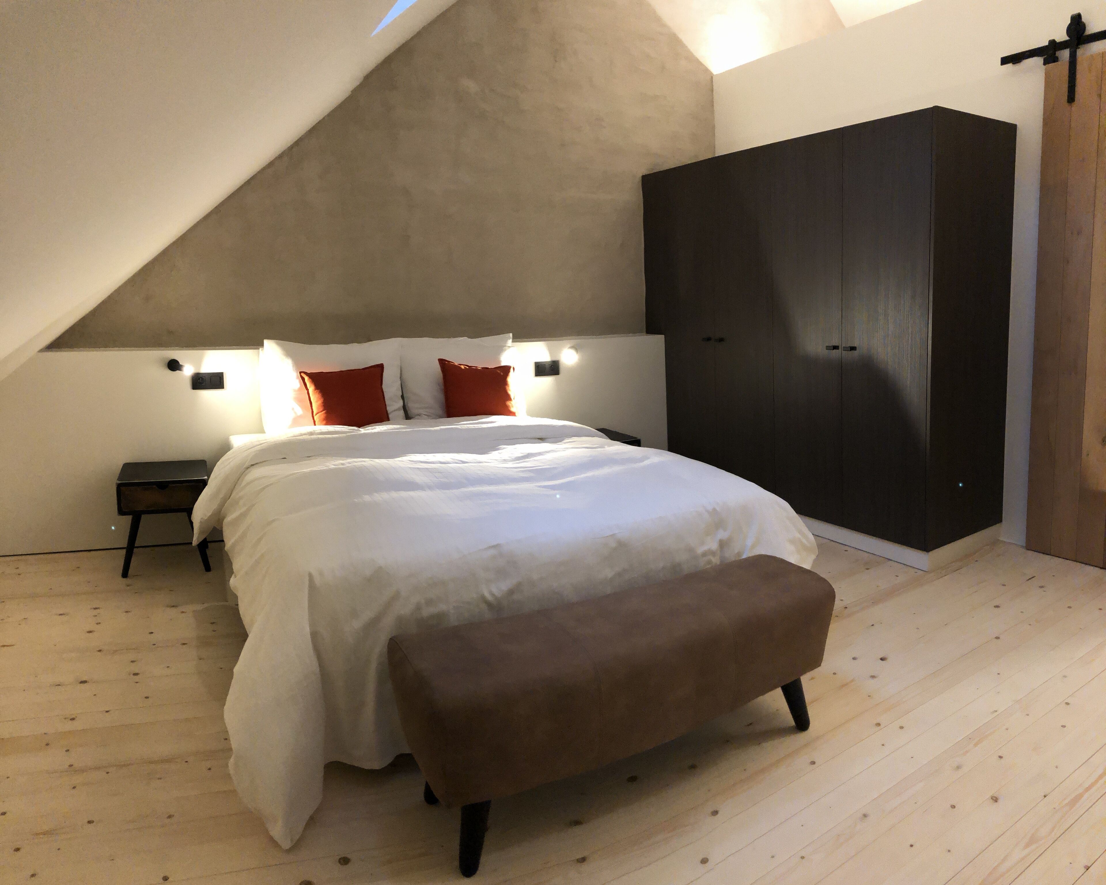 Ein Schlafzimmer mit schrägen Wänden, einem Bett mit weißer Bettwäsche und roten Kissen, einer Bank, einem dunklen Kleiderschrank und einem Nachttisch mit einer Lampe.