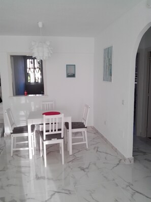 Uma área de refeições com uma mesa e cadeiras brancas, uma janela com vista e um pequeno quadro na parede.