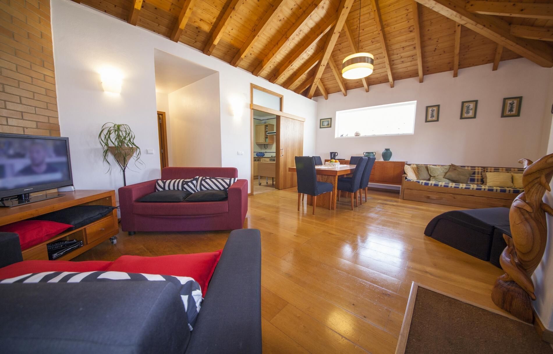 Un vaste espace de vie doté d’un plafond en bois, d’un téléviseur, d’un canapé rouge, d’une table à manger en bois avec des chaises et d’une sculpture en bois.