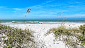 A beach with white sand, sparse vegetation, and a clear blue sky.
