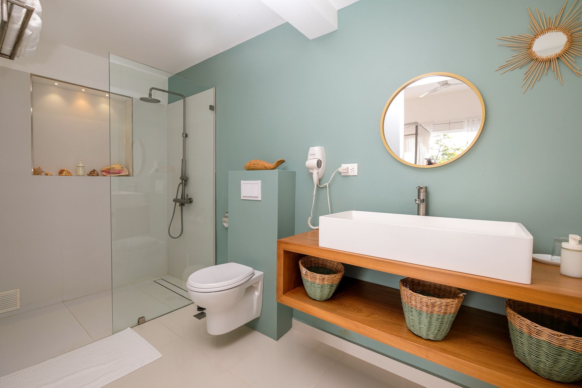 A modern bathroom with a glass shower enclosure, a white toilet, a wooden vanity with a white sink, and a round mirror.