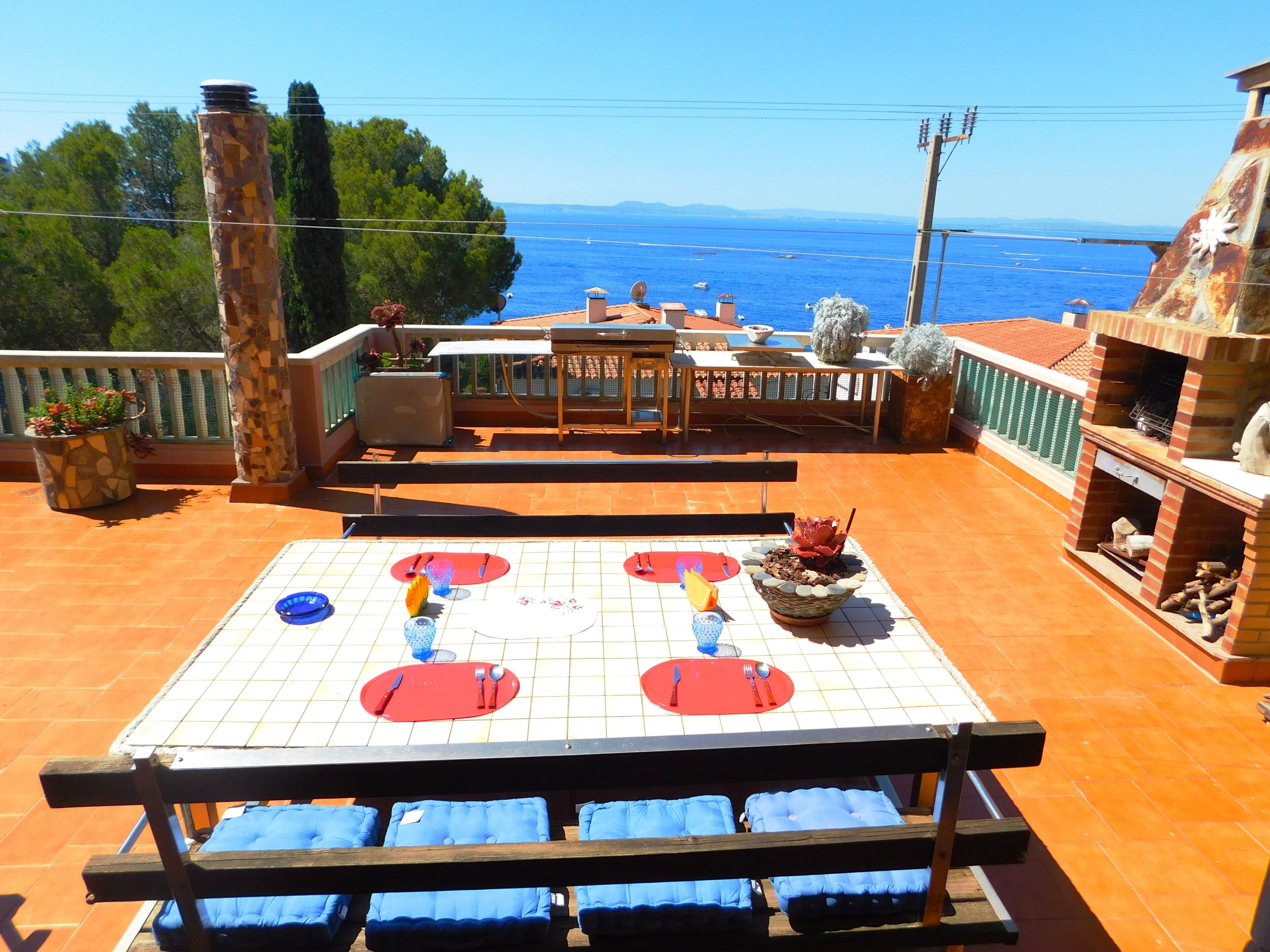 An outdoor terrace with a tiled table set for dining, blue cushioned chairs, and a view of the sea.